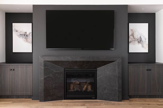 A symmetrical contemporary living room media wall featuring a large black television above a dark stone fireplace surround and inset firebox. Dark built-in cabinetry with black hardware flanks the fireplace, while abstract artwork hangs on each side. The room has a warm timber floor, charcoal walls, and recessed ceiling lighting.