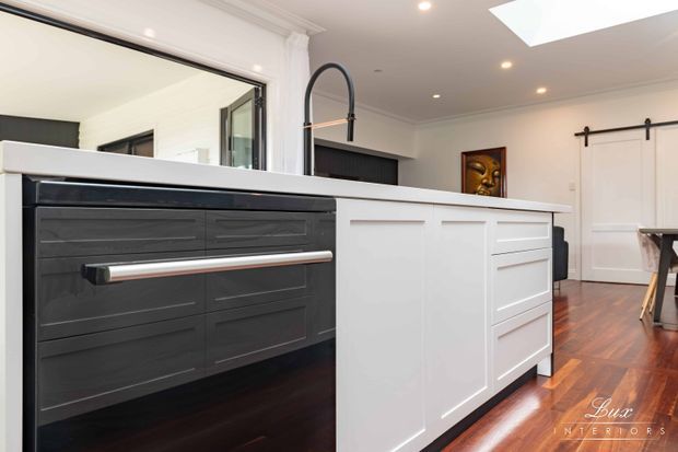 Bright contemporary kitchen centered on a large white paneled island with dark integrated cabinetry and a long metallic drawer pull. The space features dark timber flooring, white walls with decorative panel moulding, a black gooseneck faucet, skylight, recessed lighting, and a large opening to an adjoining area.
