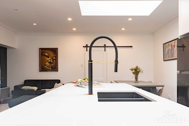 Bright contemporary kitchen viewed from behind a large white island, featuring a prominent matte-black arched gooseneck tap, integrated sink, recessed ceiling lights and a large rectangular skylight. A dining area and living room with dark sofa are visible beyond, with white cabinetry, artwork and a vase providing restrained decorative accents.