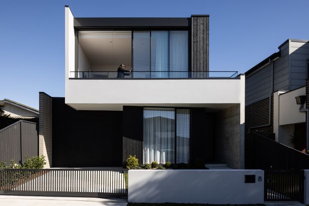 Front exterior view of a compact two-storey contemporary residence with a clean rectilinear form. The facade combines white rendered surfaces, dark vertical cladding, large glazed openings, a projecting upper-level balcony with glass balustrade, and a concealed flat roofline. A slatted front fence and landscaped entry sit in front of the house.