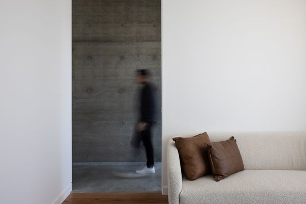A minimalist interior view featuring a textured grey concrete wall and matching polished concrete floor beyond a wide opening. A blurred person walks through the passage, while the foreground contains a cream boucle sofa with brown cushions on a warm timber floor. The restrained palette and raw concrete finish create a modern, understated atmosphere.