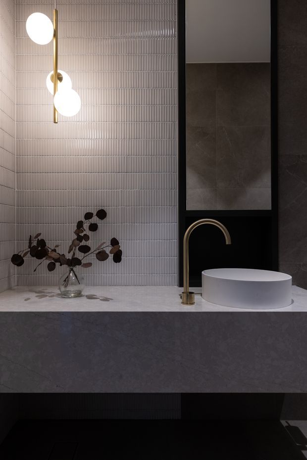A dark, refined bathroom vanity scene featuring a wall-mounted white basin on a broad grey stone-look counter, a tall brass gooseneck tap, and black-framed recessed shelving with large dark tiles. The main wall is clad in narrow vertically ribbed light-grey tiles. A sculptural globe pendant hangs above, while a small glass vase with dark foliage adds a muted organic accent.