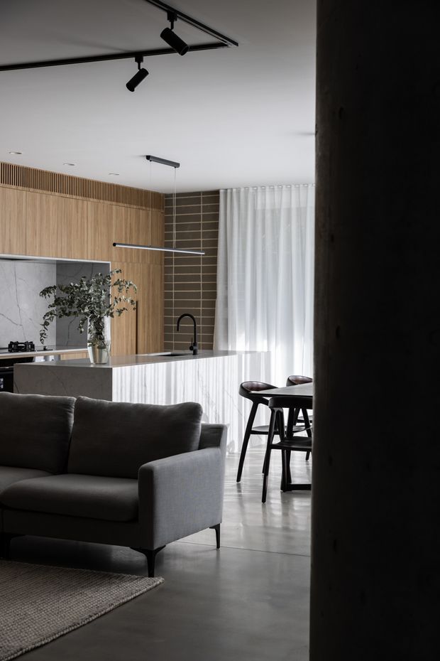 A refined open-plan kitchen and living area viewed from a darker entry or hallway. The space features warm vertical timber cabinetry and wall panelling, a pale veined stone island, dark horizontal wall tiles, sheer full-height curtains, a grey upholstered sofa, and a dark timber dining set. Black track lighting and slim pendant lights provide contemporary illumination, while polished concrete flooring and restrained furnishings create a calm, modern atmosphere.