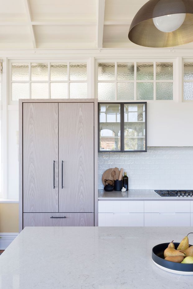 Bright, refined kitchen interior featuring a large pale engineered-stone island in the foreground, white cabinetry, a tall light timber-grain refrigerator cabinet, pale blue-grey subway tile splashback, and a black-framed internal window. White-painted timber-framed textured-glass clerestory windows and a ribbed white ceiling create an airy character-filled setting, complemented by a large bronze-toned pendant light.
