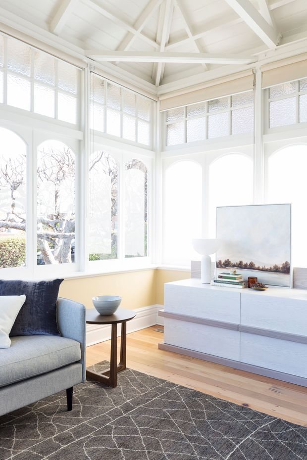 Bright, airy living room in a glazed conservatory-like space, featuring tall arched windows with textured glass, exposed white-painted roof trusses, pale yellow lower walls, light timber flooring, a grey upholstered armchair, dark patterned rug, and a low white media cabinet with artwork and table lamp.