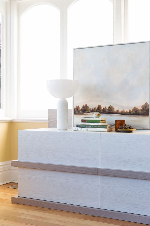 Bright, refined interior vignette featuring a large light-toned timber-finish built-in sideboard with clean slab doors and contrasting horizontal trim. A white sculptural table lamp, stacked books, decorative bowl, and large landscape artwork sit on top. Tall arched windows, white mouldings, a pale yellow wall, and timber flooring create an airy contemporary-classic setting.