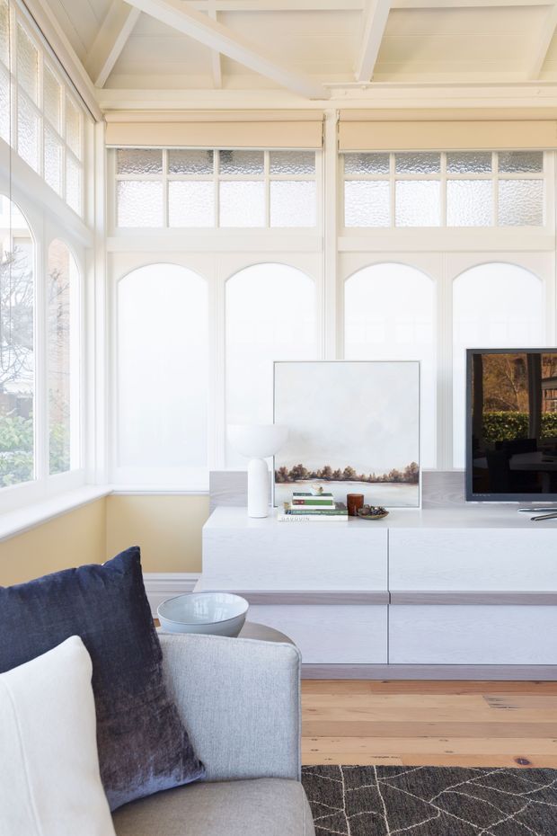 Bright, refined living room with a pale upholstered sofa in the foreground, a low white built-in media cabinet, television, decorative artwork and lamp. The room features tall arched window panels with textured glazing, extensive white-painted timber framing, and an exposed white-beamed ceiling. Light timber flooring and a dark patterned rug add contrast to the predominantly white and soft grey palette.