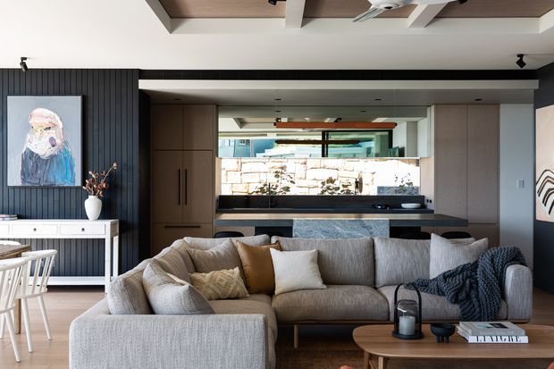 A contemporary open-plan living room with a large textured sectional sofa in the foreground, a timber coffee table, and a compact dining area to the left. Behind the living area is a kitchen with dark benchtops, timber cabinetry and a stone feature splashback, framed by dark vertical wall panelling. A layered timber ceiling detail and ceiling fan add architectural interest, while artwork and neutral furnishings create a warm, refined atmosphere.