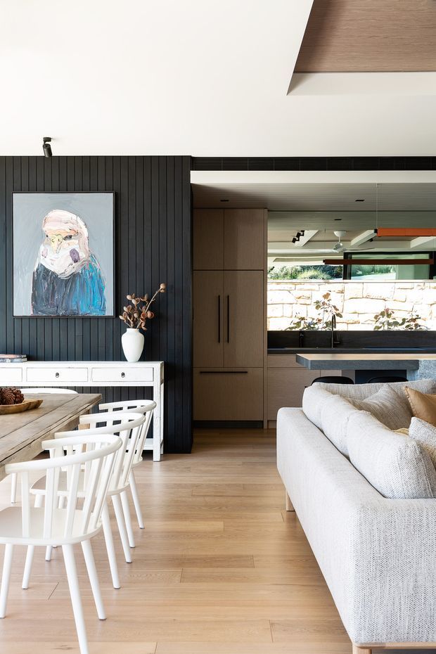 Bright open-plan residential interior combining a dining area, living room and kitchen. The space features light timber flooring and cabinetry, a dramatic black vertical timber feature wall, white dining furniture, a textured light sofa, and a dark kitchen island. A large artwork and neutral styling add visual interest, while glazing provides views toward a stone outdoor wall.