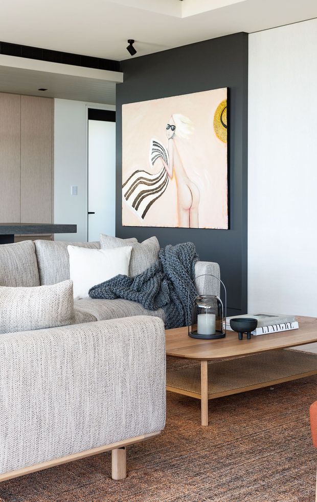 A contemporary living room with a large textured light sofa and armchair, neutral cushions, and a chunky charcoal throw arranged around a low timber coffee table. The room features a dark charcoal painted feature wall displaying large artwork, pale timber cabinetry in the background, a white ceiling with recessed detailing, black track lighting, and a warm textured rug. A partial kitchen or dining counter is visible at left.