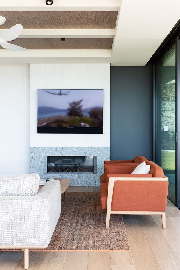 Bright contemporary living room with a wall-mounted television above a linear fireplace clad in pale blue-grey stone. The room features a white boucle sofa, terracotta upholstered timber-framed lounge chairs, a textured neutral rug, pale timber flooring, dark charcoal wall panelling, expansive black-framed glazing, and a recessed timber-look ceiling detail with a ceiling fan.