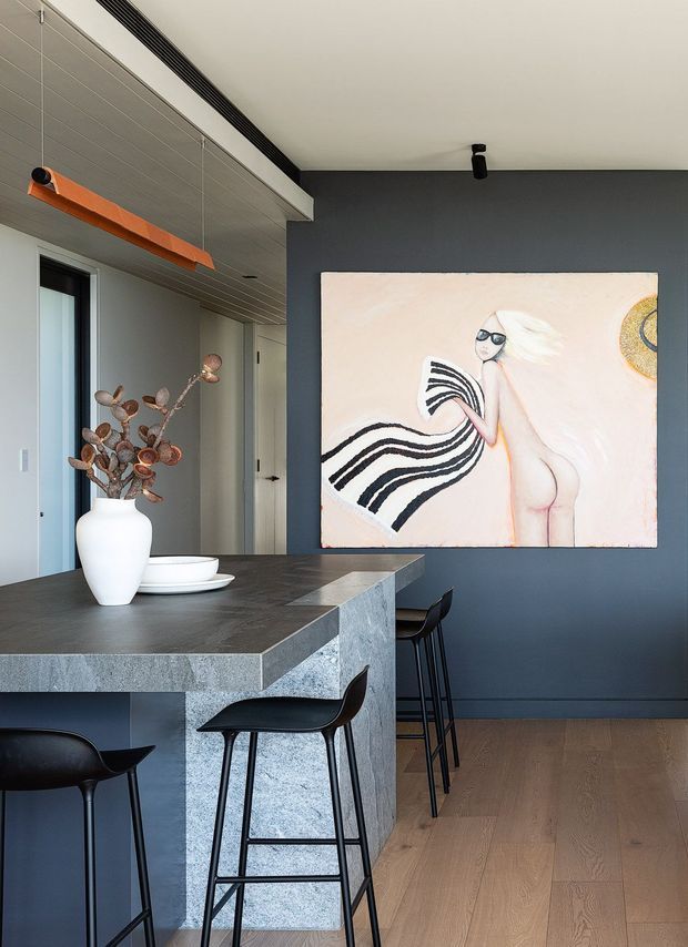 A sophisticated open-plan kitchen and dining area with a dark charcoal feature wall, large fashion artwork, grey stone island with waterfall edge, black bar stools, light timber flooring, and a sculptural orange linear pendant. The space has a refined contemporary feel with muted neutral finishes and strong graphic accents.