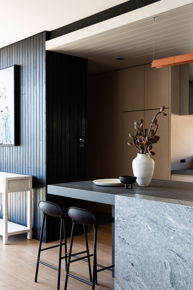 A refined open-plan kitchen and dining area with a dark vertical-panelled entry wall, warm timber cabinetry, a large grey stone island with a waterfall edge, and two black bar stools. A white vase with dried foliage decorates the island, while a linear pendant light and concealed ventilation grille add contemporary detailing.