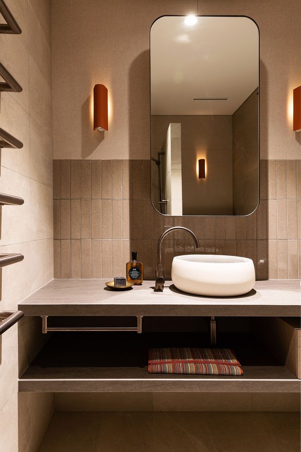 A warm, compact bathroom vanity with a wall-mounted rounded white vessel basin, brushed metal tapware, large rounded-rectangle mirror, integrated open shelving, and a textured tiled backsplash. Vertical wall sconces create a soft amber glow, while the vanity and surrounding surfaces use taupe-brown finishes.