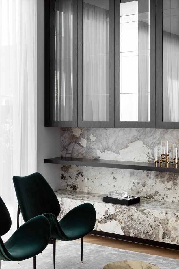 A refined interior vignette featuring dark wood-framed glass-front cabinetry above a dramatic veined stone wall and built-in console. Deep green upholstered lounge chairs sit beside a sheer-curtained window, with a dark floating shelf, decorative brass candlesticks, and a pale rug completing the sophisticated setting.