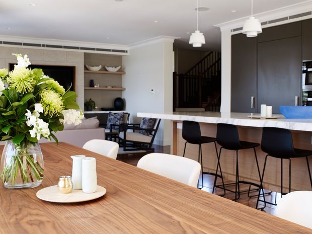Bright open-plan dining and kitchen interior with a large timber dining table in the foreground, a marble-look kitchen island with dark bar stools, dark cabinetry, pendant lights, built-in shelving, and a partially visible living area and staircase beyond. The room has a refined contemporary transitional feel with warm timber and neutral finishes.