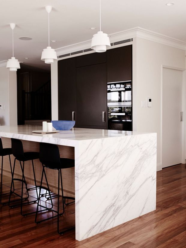 Contemporary kitchen featuring a large marble-look island with waterfall edge, integrated dark cabinetry and appliances, timber flooring, and black bar stools. White pendant lights and pale walls create a refined, high-contrast interior.