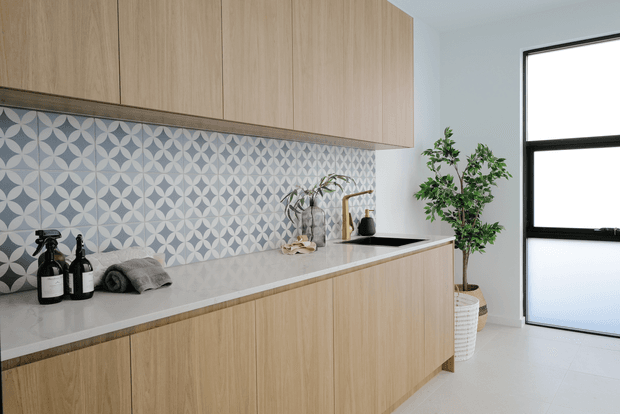 Bright contemporary kitchen with flat-panel light timber cabinetry, a pale stone benchtop, and a striking blue-grey geometric patterned tile splashback. A black sink and tap, decorative bottles, folded towels, and a potted tree are visible, with strong natural light entering through a tall black-framed window or door.