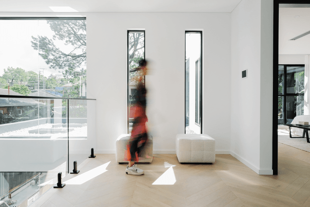 Bright, minimalist contemporary interior featuring a double-height circulation or living area with white walls, tall narrow windows, expansive glazing, and pale herringbone timber flooring. A glass balustrade overlooks a lower level, while a blurred figure stands near a low upholstered ottoman. An adjacent open room with dark-framed glazing is visible through the opening on the right.