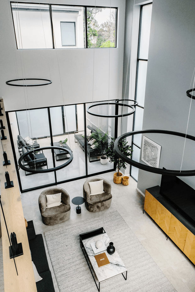 A bright, double-height contemporary living room viewed from above, featuring floor-to-ceiling black-framed glazing, multiple oversized circular pendant lights, a neutral sectional sofa and armchairs, a textured area rug, a marble-topped coffee table, indoor plants, and a black-and-timber media console. A glass-partitioned adjoining lounge area is visible below the windows.