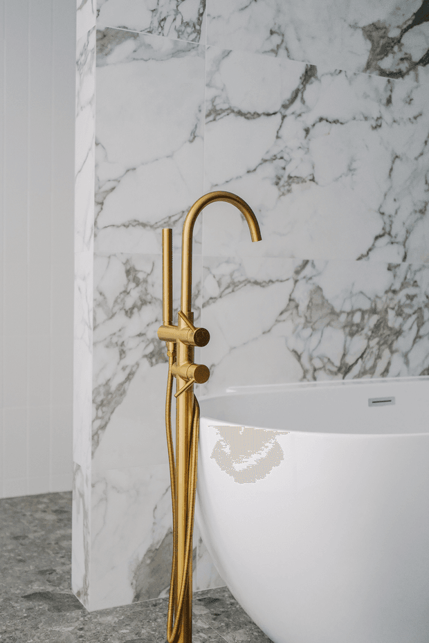 Close-up view of a luxurious contemporary bathroom featuring a freestanding white bathtub, a sculptural brushed-gold floor-mounted bath mixer with handheld shower, dramatic white marble-look wall tiling with dark veining, and grey stone-look flooring. The palette is refined and high-contrast, with gold plumbing fixtures providing a warm accent.