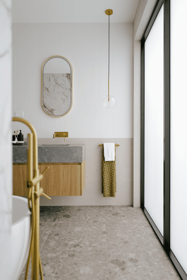 A refined contemporary bathroom with a floating natural timber vanity, grey stone benchtop, brass tapware and hardware, vertically panelled white wall tiles, and a large-format stone-look floor. An arched brass-framed mirror with a marble-like surface detail hangs above the vanity, alongside a minimalist glass pendant light and patterned towel. Full-height dark-framed glazing brings in abundant diffused light.
