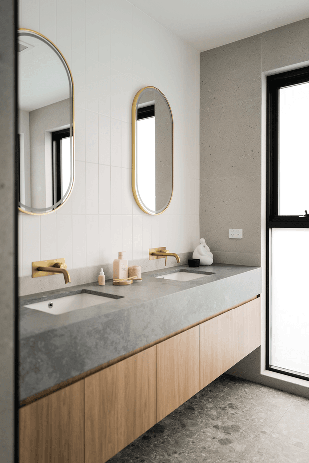 Contemporary bathroom vanity with a long grey stone benchtop, twin integrated basins, pale timber cabinetry, brushed brass wall-mounted taps, and two arched brass-framed mirrors. White tiled walls, grey stone-look wall panels, black window frames, and a dark textured floor create a refined neutral palette with warm timber and brass accents.