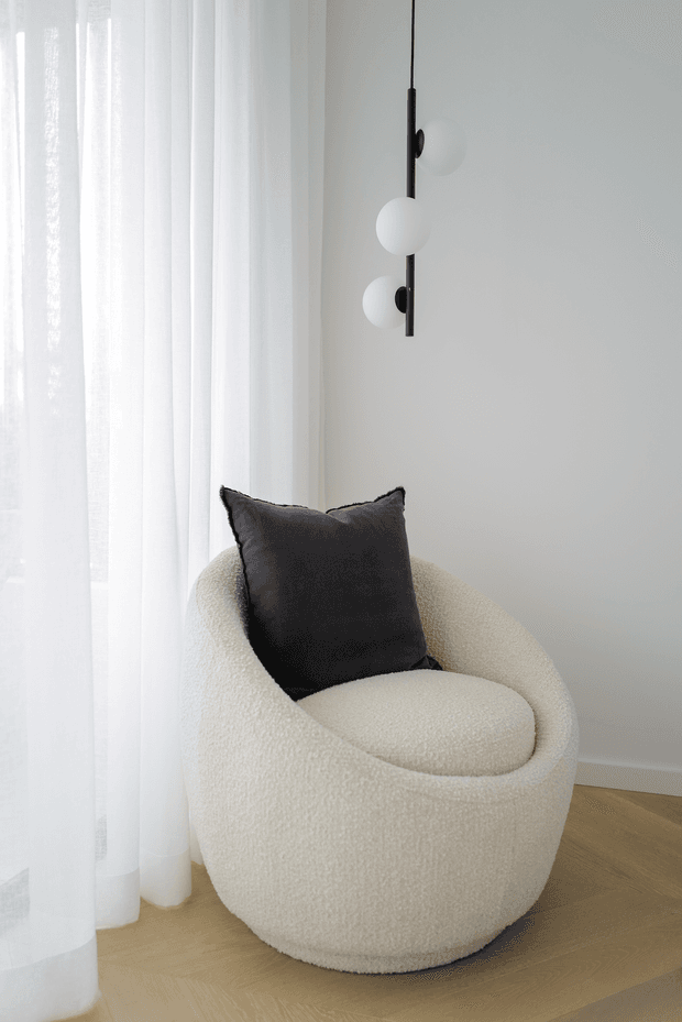 A minimalist interior corner featuring a sculptural cream bouclé lounge chair with a black cushion, positioned beside sheer white curtains. A slim black vertical pendant light with spherical white globes hangs against a pale wall, while light timber flooring adds warmth.