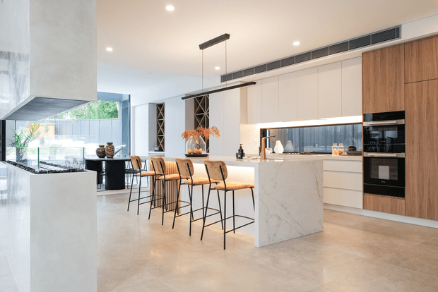A bright, spacious contemporary kitchen with a large marble-look island, waterfall benchtop, integrated cabinetry, timber pantry wall, built-in black appliances, and a row of timber-and-black bar stools. Linear pendant lights, concealed under-cabinet lighting, pale tiled flooring, and large glazing create a refined open-plan setting.