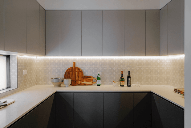 A contemporary L-shaped kitchen with matte dark lower cabinetry, taupe-grey upper cabinets, white benchtops, and a warm illuminated patterned tile splashback. The compact space includes a window, under-cabinet lighting, integrated cabinetry, and a small arrangement of cutting boards, bowls, and bottles on the counter.