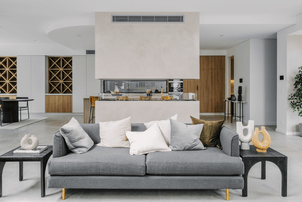 A refined open-plan living room centered on a large charcoal-grey sofa with layered neutral cushions. Behind it is a minimalist kitchen with a stone island, pale plastered feature bulkhead, timber cabinetry, integrated appliances, and a wine display. The space has light flooring, white walls and ceilings, dark furniture accents, and warm timber detailing.