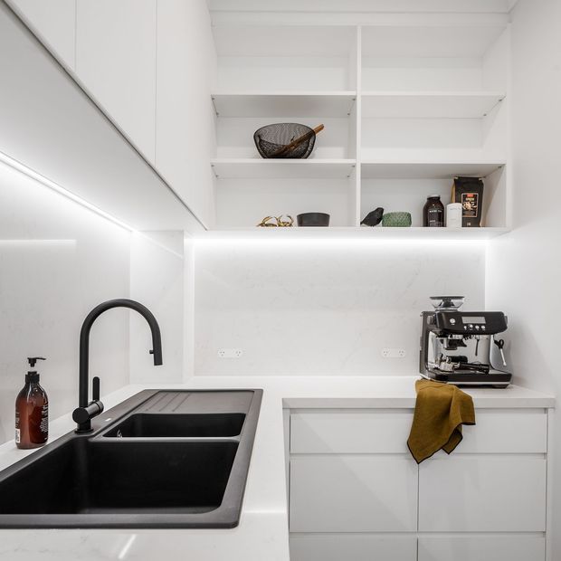 A compact, bright white kitchen or butler's pantry with streamlined flat-panel cabinetry, open shelving, integrated under-shelf lighting, a large black double-bowl sink, matte black gooseneck tapware, and a built-in coffee station. White benchtops and splashback create a clean, minimalist composition with warm mustard and dark decorative accents.