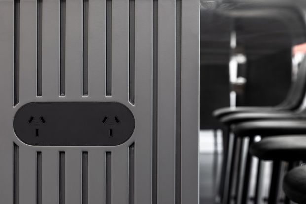 Close-up product-style image of a dark grey vertical architectural panel with integrated black electrical power outlets. Blurred black bar stools or seating are visible in the background, suggesting a commercial or hospitality interior.