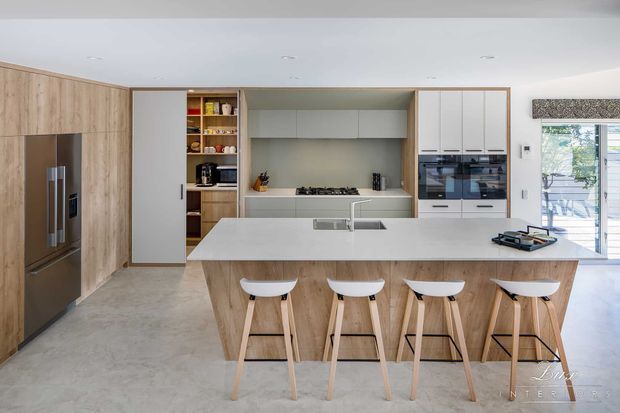 Bright contemporary kitchen with a large white island featuring an integrated sink and four light-wood bar stools. Full-height light-wood cabinetry houses a refrigerator on the left, while white cabinetry, open shelving, a cooktop, and double wall ovens line the rear wall. Pale stone-look flooring, recessed ceiling lights, and glazed doors or windows create an airy, refined space.