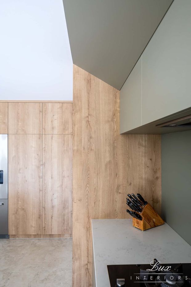 A close-up view of a contemporary kitchen featuring light timber-look cabinetry and wall panelling, a pale stone benchtop, and muted grey-green overhead cabinetry. A timber knife block sits on the benchtop, with integrated appliances partially visible. The space is clean, warm, and minimalist in appearance.