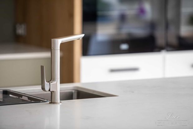 Close-up view of a contemporary kitchen sink area, featuring a tall brushed-metal single-lever tap mounted in a pale stone benchtop. A dark inset sink, cooktop edge, white cabinetry with horizontal handles, and integrated dark appliances are visible in the softly blurred background.