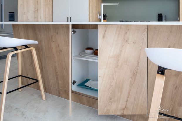 Contemporary kitchen cabinetry in a light timber finish with integrated appliances, open shelving, and a white peninsula or breakfast bar. A light-coloured stool with timber legs and black metal supports is visible, along with a pale stone or concrete-look floor. The cabinetry and clean lines create a minimalist, refined interior.