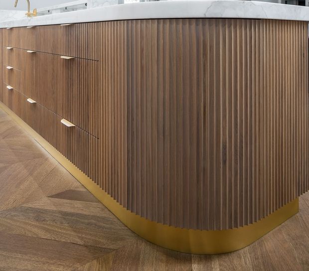 Close-up of a curved built-in kitchen island or counter with vertically fluted dark timber cabinetry, slim light-coloured drawer pulls, and a brushed brass or gold-toned plinth. The island sits on a patterned timber floor and has a pale stone benchtop with a curved edge. The image focuses on refined fixed joinery and material detailing.