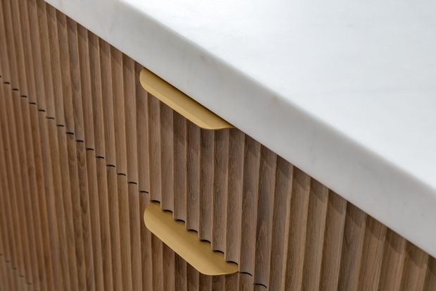 Close-up detail of vertically fluted timber cabinetry with curved brass-gold handles beneath a thick white benchtop or counter surface. The image emphasizes refined joinery, warm wood grain, rounded handle forms, and a minimalist material palette.