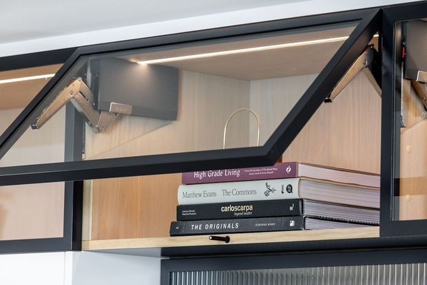 Close-up of a contemporary built-in display shelving unit with black metal-framed glass doors, warm light-wood backing, integrated spot lighting, and a small stack of books. The shelving includes angled gas-strut-style opening hardware and a curved brass-toned decorative element.