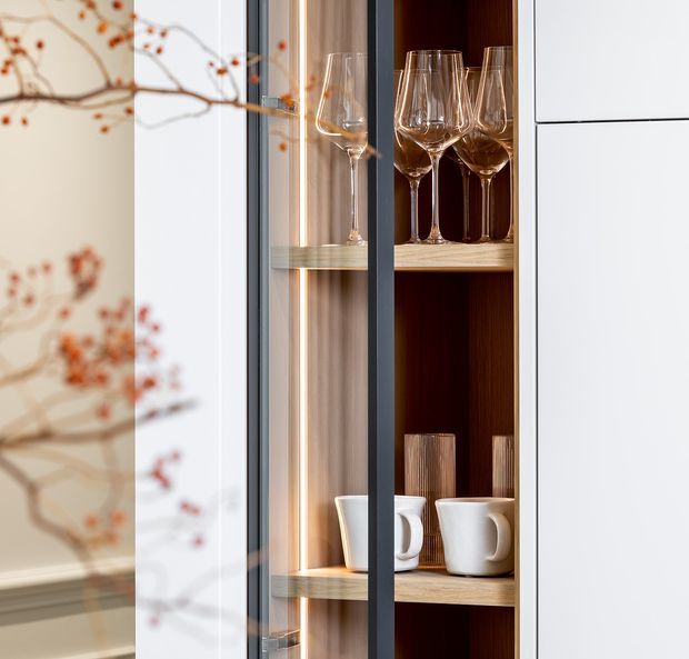 Close-up of a contemporary built-in glass-front display cabinet with dark timber interior, light timber shelves, integrated warm LED lighting, and black-framed glazing. Wine glasses and ceramic mugs are displayed inside. A softly blurred botanical patterned wall or reflection appears at the left edge.