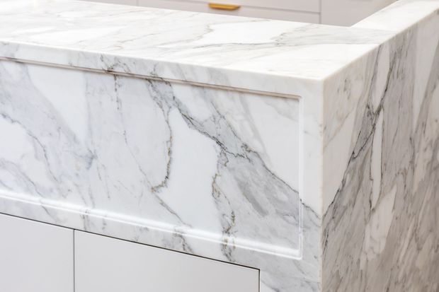 Close-up of a white marble kitchen island or counter with prominent grey veining, including a waterfall side panel and decorative framed edge detail. White cabinetry is partially visible in the background.