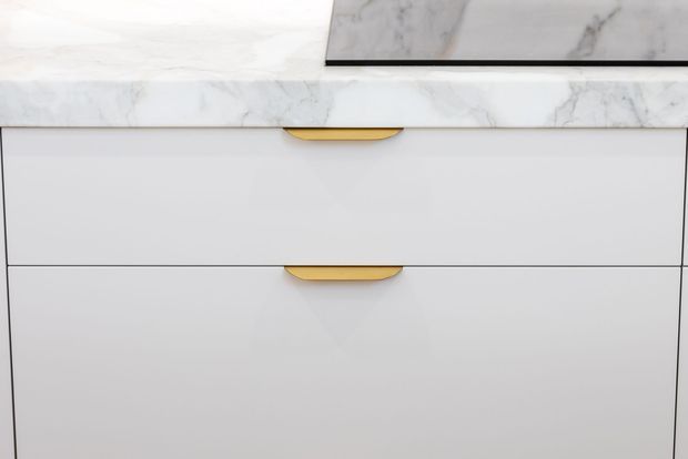 Close-up of white built-in cabinetry with clean flat drawer fronts and distinctive curved brass-gold handles. A white marble-look benchtop and matching raised backsplash or wall panel are visible above, creating a refined contemporary finish.