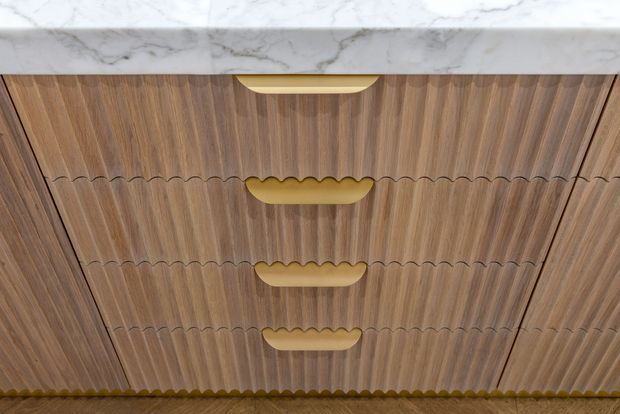 Close-up view of a decorative timber vanity or cabinetry front with vertically fluted and scalloped detailing, paired with curved gold-toned recessed pulls. A white veined marble benchtop is visible along the top edge.