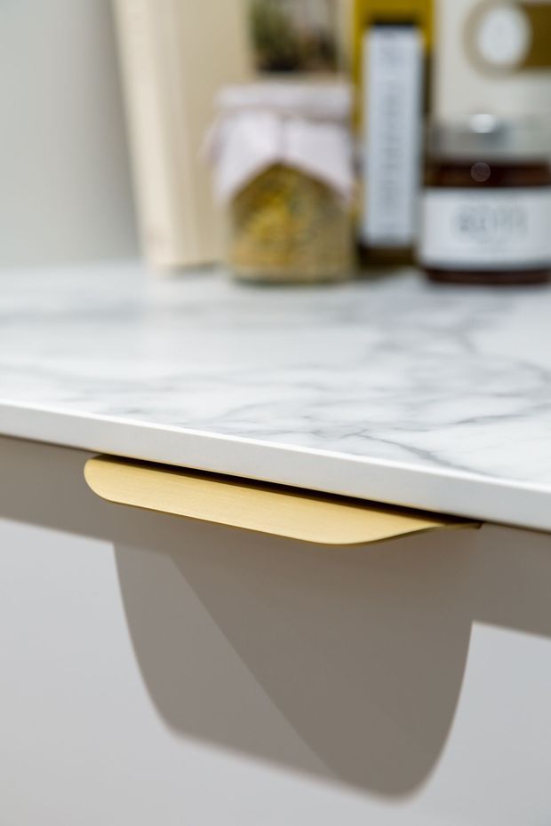 Close-up detail of a light-coloured built-in cabinet or vanity with a white marble-look benchtop and a prominent curved brushed-gold handle. Blurred toiletry or cosmetic containers sit on the countertop, suggesting a bathroom or dressing area. The image focuses on the cabinetry hardware and stone surface rather than the wider room.
