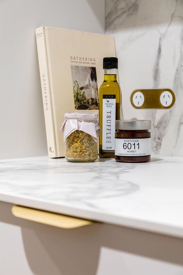 Close-up interior detail of a refined kitchen or pantry setting, featuring a white marble-look benchtop and matching marble splashback, warm neutral cabinetry with a slim recessed handle, and a small styled arrangement of a cookbook, truffle oil, honey jar and pantry ingredients. A double power outlet is integrated into the splashback.