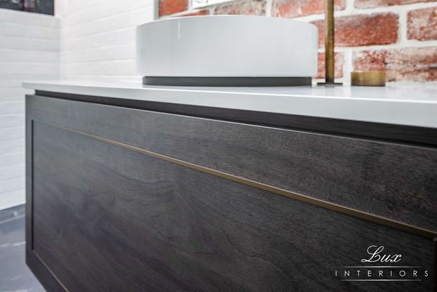 Close-up of a contemporary dark timber vanity or built-in cabinet with a slim brass-gold horizontal inlay handle and a light stone benchtop. The cabinet is set against white-painted brick and exposed red brick walls, with a rounded white vessel basin and dark tap visible on top.