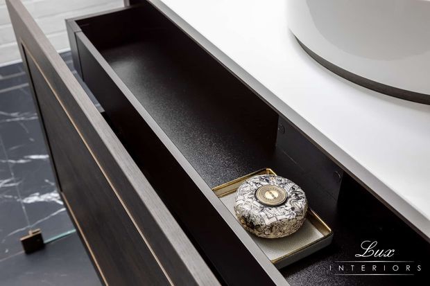 Close-up of a sleek dark timber vanity drawer with a black interior and a small decorative tray, positioned beneath a white curved benchtop or basin. Grey-toned flooring and refined detailing create a contemporary bathroom joinery setting.