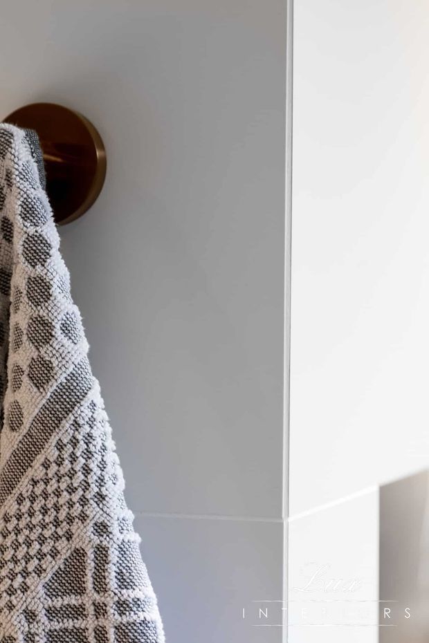 Close-up architectural detail of a pale grey tiled wall with a circular bronze towel hook and a textured black-and-white patterned towel hanging from it. A bright white adjoining wall or shower edge is visible at right, creating a clean, refined bathroom setting.