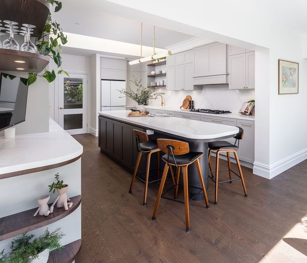 Bright contemporary kitchen with a large island, dark fixed cabinetry, white cabinetry and benchtops, integrated appliances, timber flooring, and a small breakfast-bar seating area. Open shelving and curved built-in display joinery connect the kitchen to the adjacent living space.